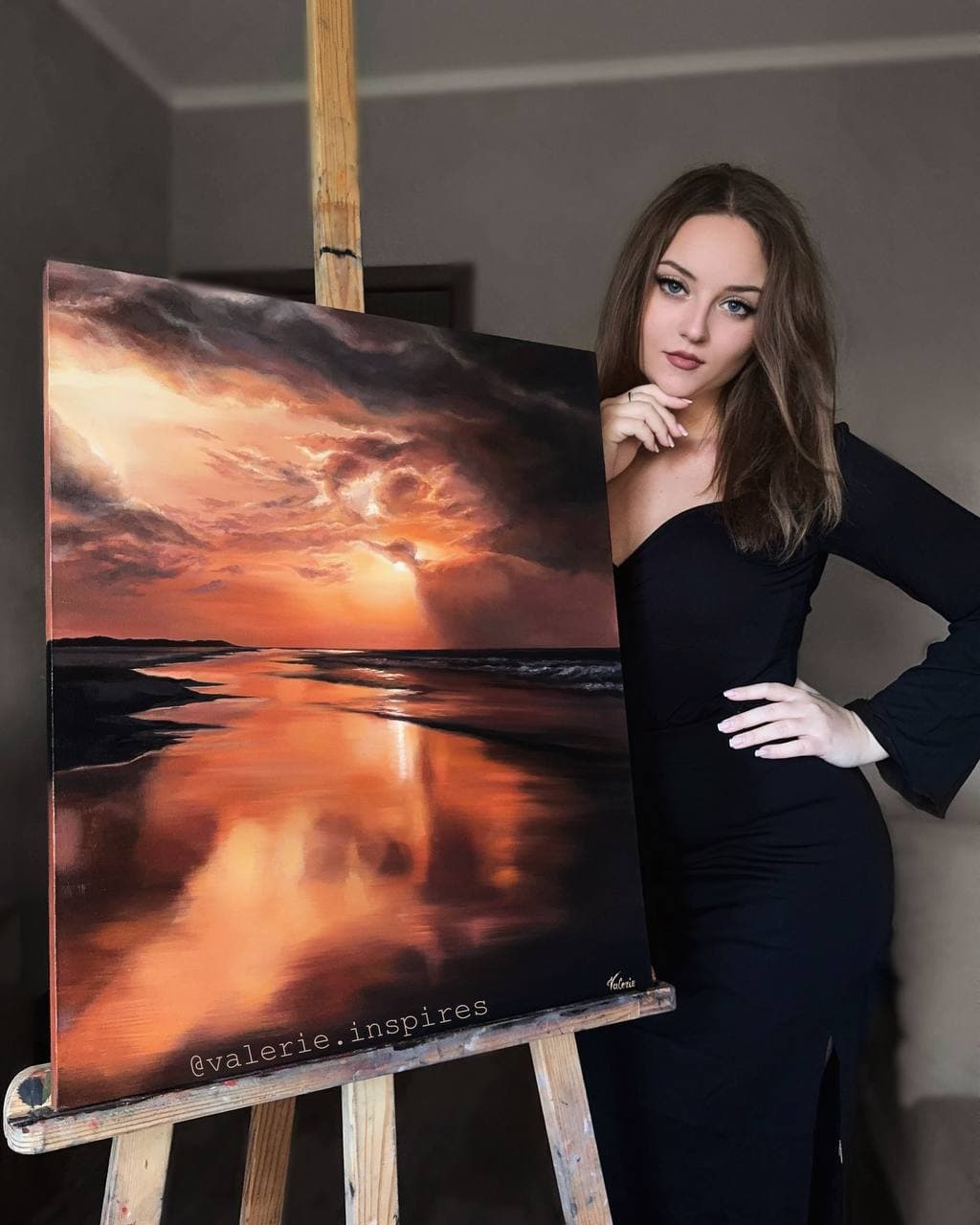 Valerie beside her oil painting of an orange sunset reflected on a quiet shoreline.