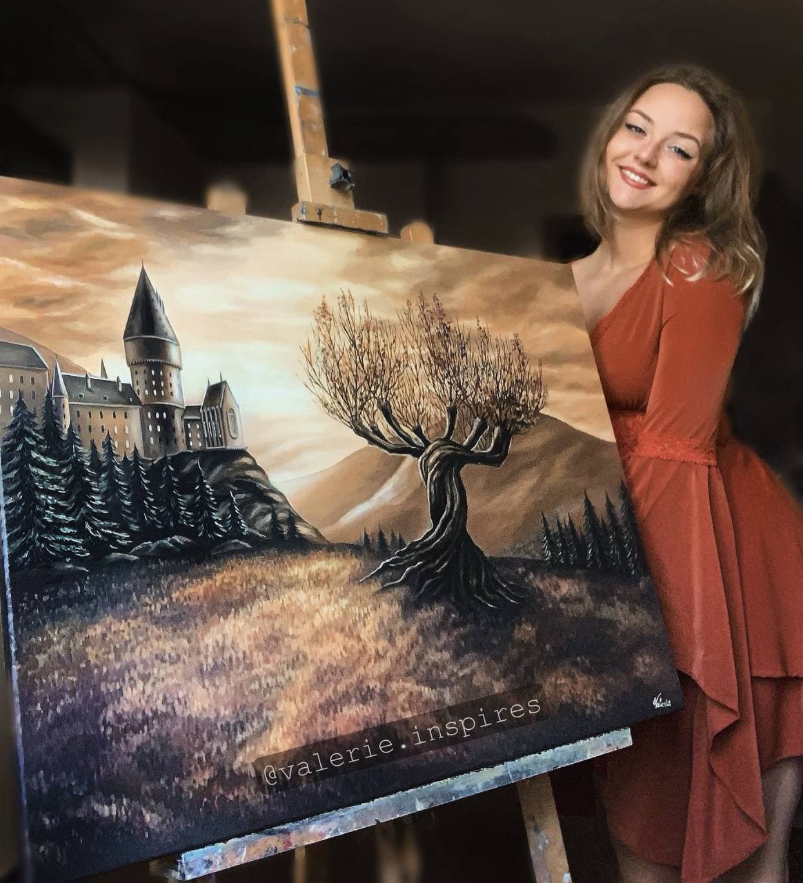 Valerie beside her oil painting of a castle, autumn hills and a twisting tree.