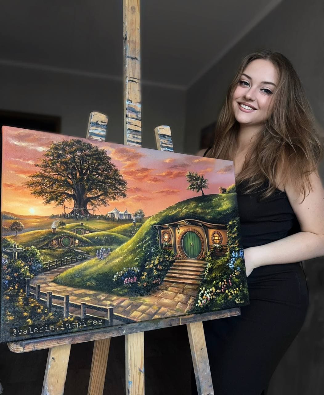Valerie beside her oil painting of green hills, round-doored cottages and a large tree at sunset.